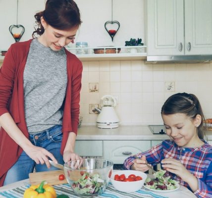 Happy mother and cheerful daughter cook salad together in the kitchen at home cutting vegetables and talking. Family, cooking, and people concept