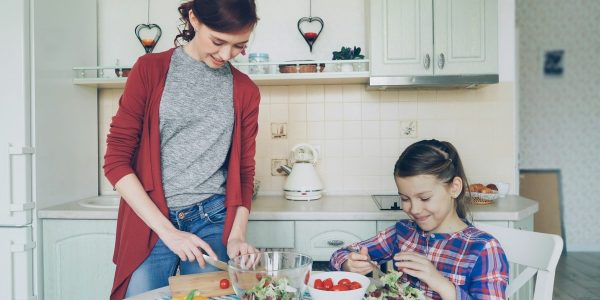 Happy mother and cheerful daughter cook salad together in the kitchen at home cutting vegetables and talking. Family, cooking, and people concept